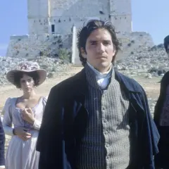 Młody mężczyzna w stroju z epoki, jak z filmu hrabia Monte Cristo 2024, stoi na tle kamiennej budowli. Obok niego kobieta w kapeluszu i inny mężczyzna.