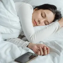 Mujer duerme profundamente, como si soñara que no puede abrir los ojos. Un teléfono móvil descansa cerca.