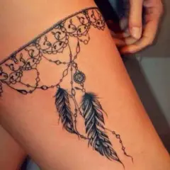 Delikatny tatuaż podwiązka z koronkowym wzorem i wiszącymi piórkami na udzie.