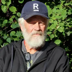 En äldre man med vitt skägg och keps med ett R på sitter utomhus. Han är en del av Ronny Eriksson familj.