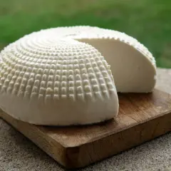 Formaggio fresco a forma di cupola, con una texture a griglia, su un tagliere di legno. Una fetta è stata rimossa, rivelando la morbida ricotta vaccina.