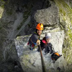 Uśmiechnięci alpiniści na skalnej półce, gotowi na wejście na mnicha. Widok z góry na stromą ścianę.