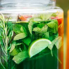 Zielony, orzeźwiający syrop z mięty z dodatkiem limonki i rozmarynu, w dużym słoiku. Obok pomarańczowy napój.