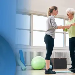 Physiotherapeutin hilft älterer Frau auf Balance Board, um Sturzgefahr zu reduzieren.