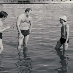 Trzech mężczyzn w kąpielówkach stoi w wodzie. Jeden z nich, Stanisław Tym w filmie Rejs, rozmawia z kolegami.