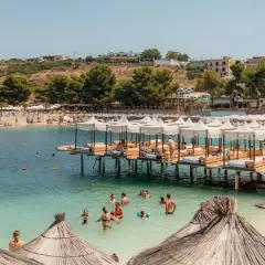 Słoneczny dzień na plaży Ksamil. Ludzie kąpią się w turkusowej wodzie, odpoczywają pod białymi parasolami na pomostach i na piaszczystym brzegu.