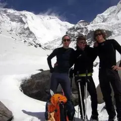 Tres montañistas posan con equipo de esquí y mochila frente a un paisaje nevado y montañoso.