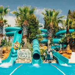 Vattenrutschkanor i Waterworld Ayia Napa med palmer och sol. Två personer åker i en ring nerför en blå vattenrutschkana.
