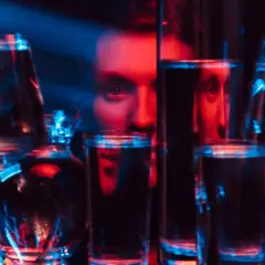 Twarz mężczyzny wyłania się zza kieliszków i szklanek, sugerując ukryty alkoholizm objawy. Czerwono-niebieskie światło podkreśla niepokój.