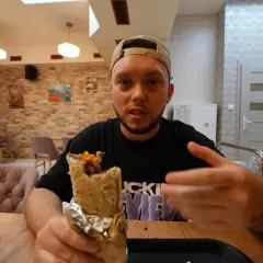 Mężczyzna w czapce z daszkiem trzyma w ręku dużego kebaba, wyglądając jak prawdziwy lider kebab. W tle widać wnętrze restauracji.