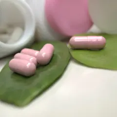 Różowe kapsułki, być może zawierające ketoprofen, leżą na zielonych liściach obok otwartych pojemników z lekami.