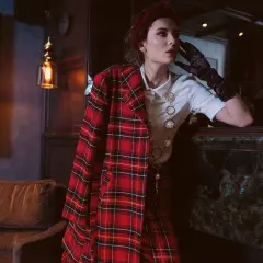 Une femme pose, vêtue d'un blazer au **motif tartan** rouge et noir, avec un collier perlé.