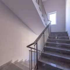 Nowoczesne schody w bloku z szarymi stopniami i metalową balustradą. Jasne ściany i okno zapewniają dobre oświetlenie, tworząc przyjazną atmosferę. Ogrzewanie klatki schodowej w bloku zapewnia komfort.