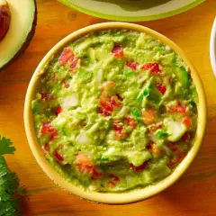 Miseczka guacamole z pomidorami i cebulą, obok awokado, kolendra i plasterki jalapeno.