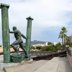 Statua di Ercole che sorregge le colonne d'Ercole oggi, con palme e edifici sullo sfondo.