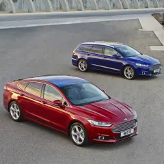 Dwa samochody Ford MK4: czerwony sedan i niebieskie kombi, stoją na asfaltowej nawierzchni przy betonowej konstrukcji i morzu.