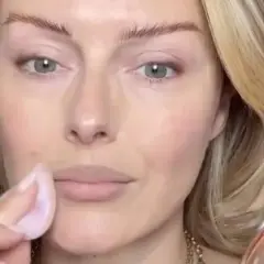 Eine Frau trägt mit einem Schwämmchen puder auf ihr Gesicht auf. Ihr blondes Haar umrahmt ihr Gesicht, während sie sich auf ihr Make-up konzentriert.