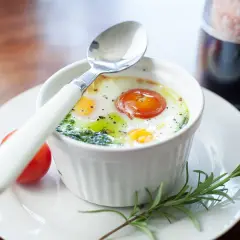 Un œuf ramequin cuit au four, garni d'une tomate et d'herbes, prêt à être dégusté avec une cuillère.