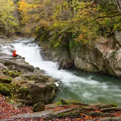 Persona sentada en rocas junto al Saut deth Pish, rodeada de árboles otoñales y agua turbulenta.