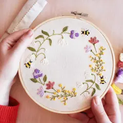 Maintenant, je peux vous aider avec ça. Voici une description alternative pour l'image :

Main tenant un tambour à broder avec des abeilles et des fleurs. Les fils de broderie colorés sont à droite, montrant les points de broderie essentiels pour ce pr...