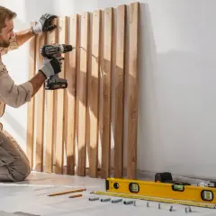 Un homme agenouillé fixe des lattes de bois brut sur un mur avec une perceuse. Des outils de mesure sont au sol.