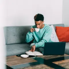 Ung man sitter i soffan med en bok och en kopp kaffe. Han tar en klunk tein koffein medan han läser.