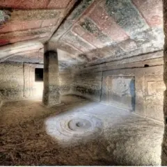 Interno di una tomba etrusca con soffitto dipinto e un pilastro centrale. Un cerchio inciso sul pavimento sembra un antico incantesimo putifigari.
