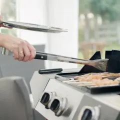Person i blå klänning använder tång för att vända kycklingspett på en fyrkantig kolgrill.