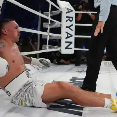 Boxer Oleksandr Usyk sits on the canvas, leaning against the ropes, appearing to recover from a usyk low blow. The referee stands nearby.