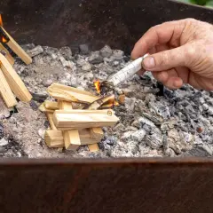 Maintenant, comment allumer un barbecue au charbon : une main allume un morceau de papier roulé pour enflammer des brindilles dans le brasero.