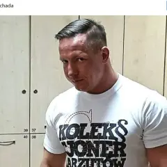 Tomasz Chada w białej koszulce z napisem "KOLEKS JONER ARZUTOW", pozuje na tle szafek.
