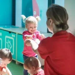 Nauczycielka w czerwonej bluzce bawi się z dziećmi w różowych i czerwonych ubrankach.