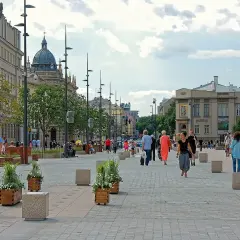 Ludzie spacerują po pięknym, wybrukowanym placu Litewskim, otoczonym zabytkowymi budynkami i zielenią.