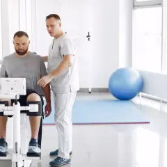 Fizjoterapeuta pomaga pacjentowi na maszynie rehabilitacyjnej w jasnym gabinecie.