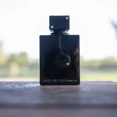 Elegancka czarna butelka perfum Club de Nuit Intense Man, ozdobiona łańcuszkiem i dyskiem z logo, stoi na drewnianym stole.