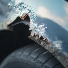 Detaljerad bild av rostig bilskärm. En del av skärmkanten har flagnat och blottar metallen under, redo att laga rost bil. Däcket syns nedtill.