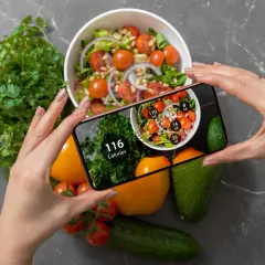 Ręce trzymają smartfon z aplikacją pokazującą kalorie. Na ekranie widać sałatkę i zioła, a obok na blacie leżą warzywa. Zastanawiasz się, ile kalorii żeby przytyć?