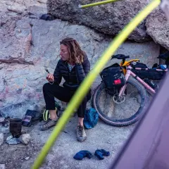 Pause sur une sortie gravel : une cycliste se repose près de son vélo rose, une tasse à la main, au milieu d'un paysage rocheux.
