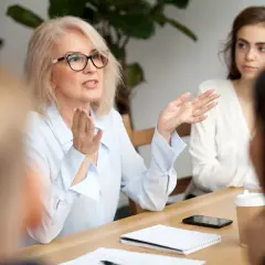 Podczas rady pedagogicznej, doświadczona nauczycielka z okularami energicznie tłumaczy coś koleżankom przy stole.