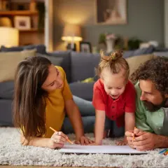 Rodzina rysuje kredkami na kartce papieru leżąc na dywanie w salonie.