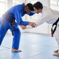 Dwóch mężczyzn w kimonach do BJJ, jeden w niebieskim z brązowym pasem, drugi w białym z czarnym pasem, ćwiczy chwyty na macie.