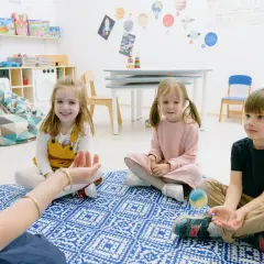 Dzieci biorą udział w zabawach integracyjnych w przedszkolu, siedząc na dywanie i słuchając nauczycielki.