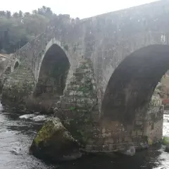 El histórico puente Maceira cruza un río caudaloso, con casas de tejados rojos y árboles frondosos al fondo.