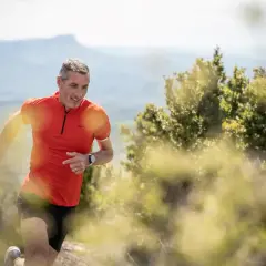 Un homme en tenue de trail run, souriant, court sur un sentier de montagne.