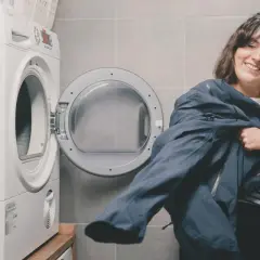 Uśmiechnięta kobieta trzyma granatową kurtkę, przygotowując się do prania jej zgodnie z instrukcją, jak prać Gore-Tex, obok otwartej pralki.