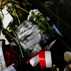 Czarne tło, bukiety czerwonych róż, białe kwiaty, zdjęcie mężczyzny w okularach, polska flaga. W tym miejscu panuje cisza.