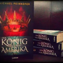 Michael Peinkofer präsentiert "Der König von Amerika". Ein Buchcover mit Krone und gestapelte Exemplare des Romans.