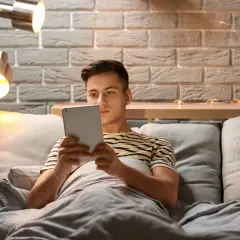 Młody mężczyzna leży w łóżku, pogrążony w tablecie. Czy to początek uzależnienia od masturbacji, czy tylko chwila relaksu?