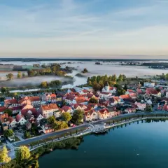Poranna mgła nad rzeką i polami, otaczająca urokliwe miasteczko z czerwonymi dachami. To prawdziwa kraina 100 jezior, gdzie spokój miesza się z pięknem natury.