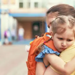 Mała dziewczynka z pomarańczowym plecakiem przytula się do mamy. To jej pierwszy dzień w przedszkolu, widać niepewność na jej twarzy.